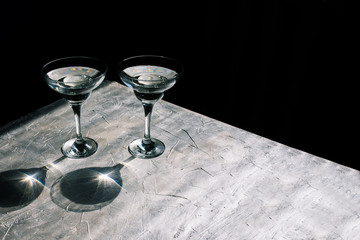 Two cocktail glasses on a silver table and black background. Glass goblets and reflection from sunlight. Glare and natural shadows.