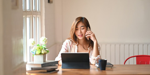 Photo a beautiful woman using a smartphone to calling someone while sitting in front a computer tablet with keyboard case at wooden working table over comfortable living room as background.