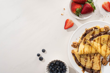 Wall Mural - top view of tasty crepes with chocolate spread and walnuts on plate near bowls with blueberries and strawberries on grey background