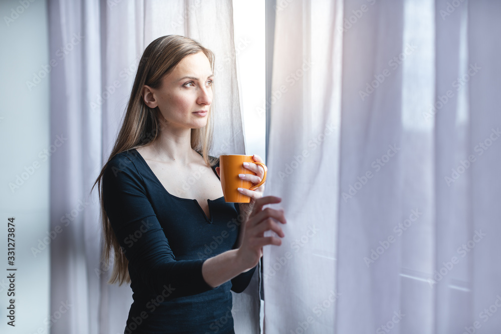 Frau steht mit Kaffee allein am Fenster während der Covid-19 Ausgangssperre - obrazy, fototapety, plakaty 