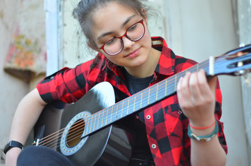 A young girl with glasses and her hair in a bun plays a classic black guitar sitting on the porch of an old house. The girl is wearing a red-and-black checked shirt and black jeans. Selective focus.