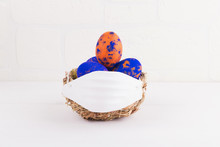 Easter Time In Quarantine Concept. Face Mask On A Nest With With Classic Blue And Orange Speckled Easter Eggs On The Table On A White Breakwall Background