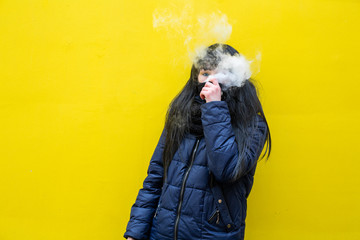 Wall Mural - Vape teenager. Young pretty white caucasian brunette girl with blue eyes and in a blue jacket smoking an electronic cigarette opposite yellow wall on the street in spring. Virus protection. Bad habit.