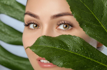 Beautiful woman with clean skin on a background of a sheet of monstera with drops of water. Eyelash Extension Procedure.