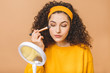 © denis_vermenko - Perfect view. Morning routine. Joyful young curly woman is sapplying foundation cream on her skin. She is looking at mirror in her hand. Isolated on beige background