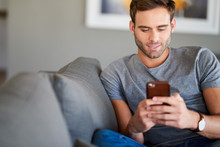 Smiling Young Man Texting On His Living Room Sofa