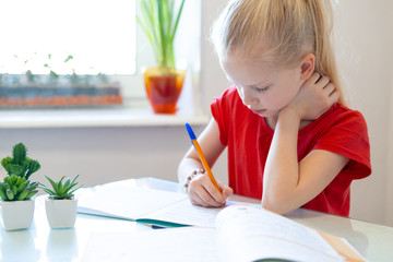 Wall Mural - Serios blonde schoolgirl sitting at home and doing school homework, writing in notebook. Training books on table. Back to school. Distance learning online education