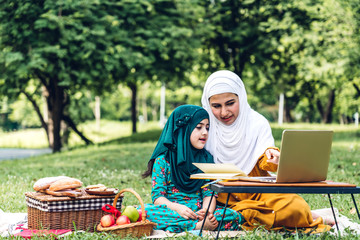 Wall Mural - Portrait of happy religious enjoy happy love asian family arabic muslim mother and little muslim girls child with hijab dress smiling and having fun looking at laptop computer in summer park