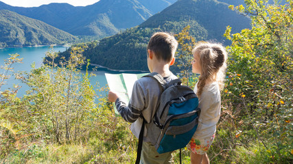 Independent little children scouts with tourist backpacks stand on the shore of a mountain lake in the Alps and pave the way for a family boy scout weekend camping in the forest.