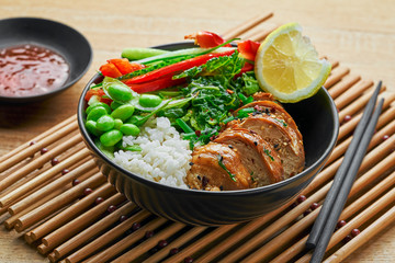 Buddha bowl meal with teriyaki chicken fillet, fresh vegetable salad, stew cabbage, edamame and basmati rice on a wooden table. Delicious dish in a black bowl.