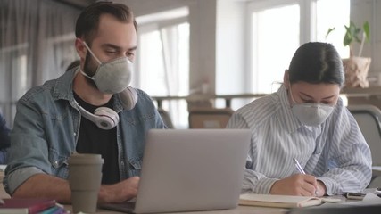 Canvas Print - Young concentrated business people colleagues indoors in office using laptop computer writing notes in notebook wearing masks because of coronavirus