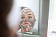 © H_Ko - Close-up view of beautiful young woman applying clay mask on face. Female looking in small mirror. Moisturizing treatment. Morning ritual and skincare procedure concept