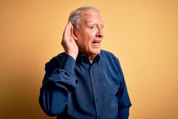 Canvas Print - Grey haired senior man wearing casual blue shirt standing over yellow background smiling with hand over ear listening an hearing to rumor or gossip. Deafness concept.