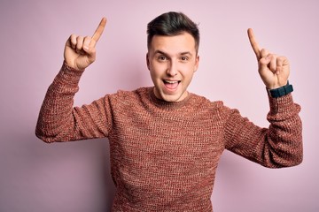 Young handsome caucasian man wearing casual winter sweater over pink isolated background smiling amazed and surprised and pointing up with fingers and raised arms.