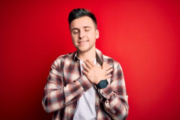 Young handsome caucasian man wearing casual modern shirt over red isolated background smiling with hands on chest with closed eyes and grateful gesture on face. Health concept.