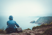 Woman With Raincoat And Backpack Sitting On The Edge Of A Cliff Watching The Sea On A Rainy And Moody Day. Outdoors Conceptual Toned Image With Copy Space.