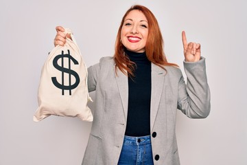 Canvas Print - Young beautiful redhead woman holding bag with money and dollar sign over white background surprised with an idea or question pointing finger with happy face, number one