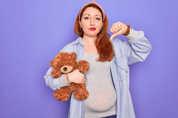 Poster - Young redhead pregnant woman expecting baby holding teddy bear over purple background with angry face, negative sign showing dislike with thumbs down, rejection concept