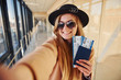 © standret - Young female passenger in warm clothes showing tickets in airport hall