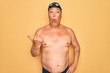 © Krakenimages.com - Middle age senior grey-haired swimmer man wearing swimsuit, cap and goggles Surprised pointing with hand finger to the side, open mouth amazed expression.