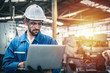 © Pitchayaarch - Confident engineer in blue jumpsuit holding laptop computer in a warehouse.