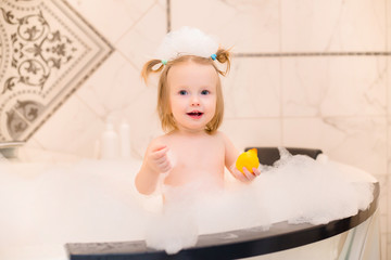 Wall Mural - Toddler girl bathes in bath with foam at home, plays with rubber duck