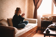Young Thoughtful Woman Drinking Tea And Eating Chocolate While Looking Through Window Sitting On Sofa In Living Room At Home