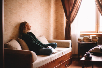 Wall Mural - Young upset woman holding book looking through window while sitting on sofa in living room at home