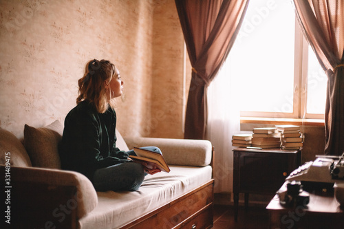 Fotomural Young thoughtful woman holding book looking through window while sitting on sofa