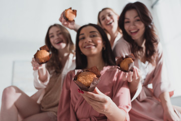 Wall Mural - Selective focus of multicultural women showing muffins, smiling and looking at camera at bachelorette party