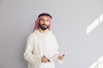 Wall Mural - Attractive smiling arab man writes in clipboard on gray background