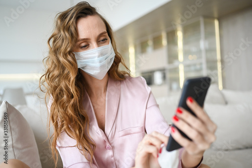 European woman in face mask cleaning the phone by hand sanitizer, using cotton wool with alcohol to wipe to avoid contaminating with Corona virus. Cleaning mobile phone to eliminate germs, Covid-19.