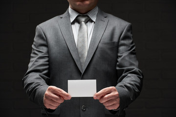 white blank business card closeup in businessman hand, gray suit, dark wall background