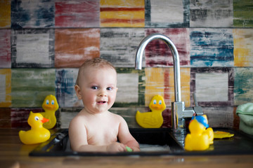 Wall Mural - Cute snmiling baby taking bath in kitchen sink. Child playing with foam and soap bubbles in sunny kitchen with rubber ducks, little boy bathing