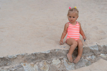 Sad cute caucasian small girl in pink swimming suit sits on sand, she is upset and ready to cry because of bathing ban cause of koronavirus