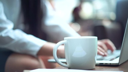 Ceramic coffee cup foreground and business female hands typing computer notebook keyboard background