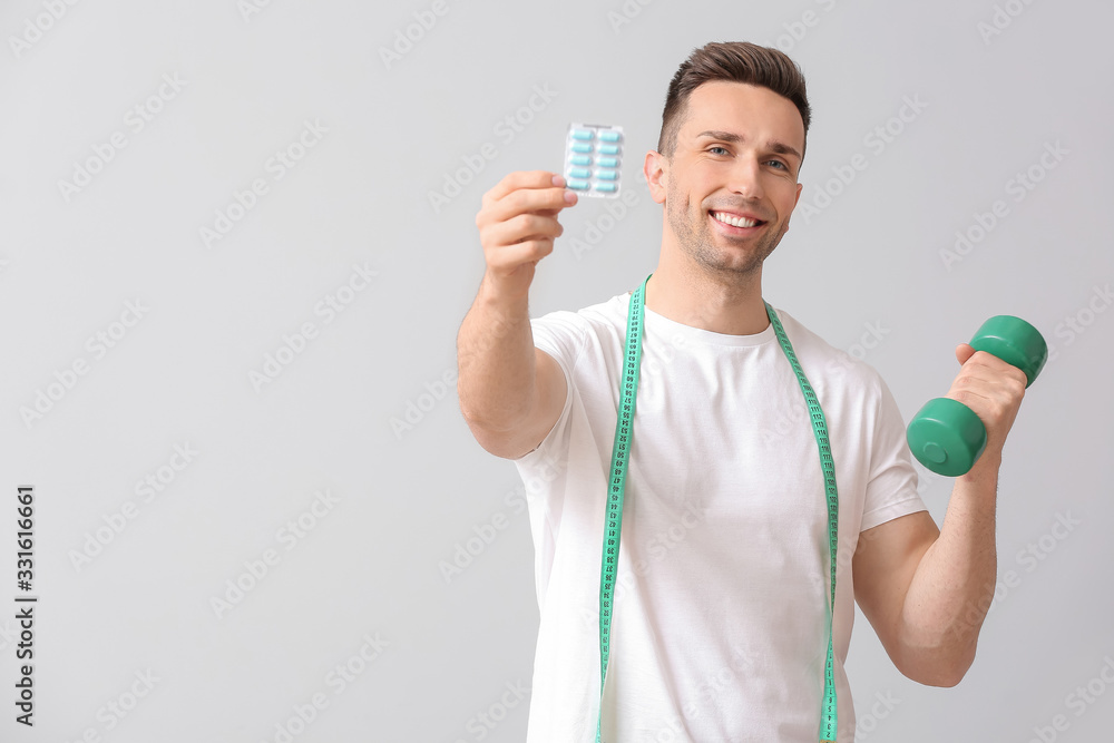 Young man with weight loss pills and dumbbell on light background