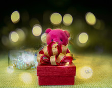 A Red Gift Box With Golden Ribbon In A Pink Teddy Bear Arms Standing On Green Fabric,  A Glass Bottle In The Back, Under Bokeh Yellow Lighting On Dark Background