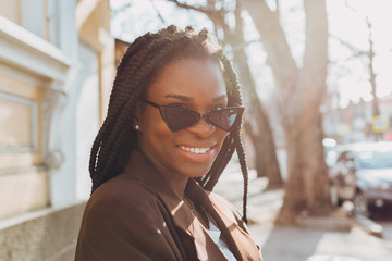 Wall Mural - Close up portrait of a beautiful young african american woman with pigtails hairstyle in a brown business suit walks along spring streets