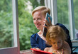 Mother and daughter by the open window, mother communicates cheerfully using a tablet, holds it near her ear like a telephone, daughter sits on her mother and examines her own hands.