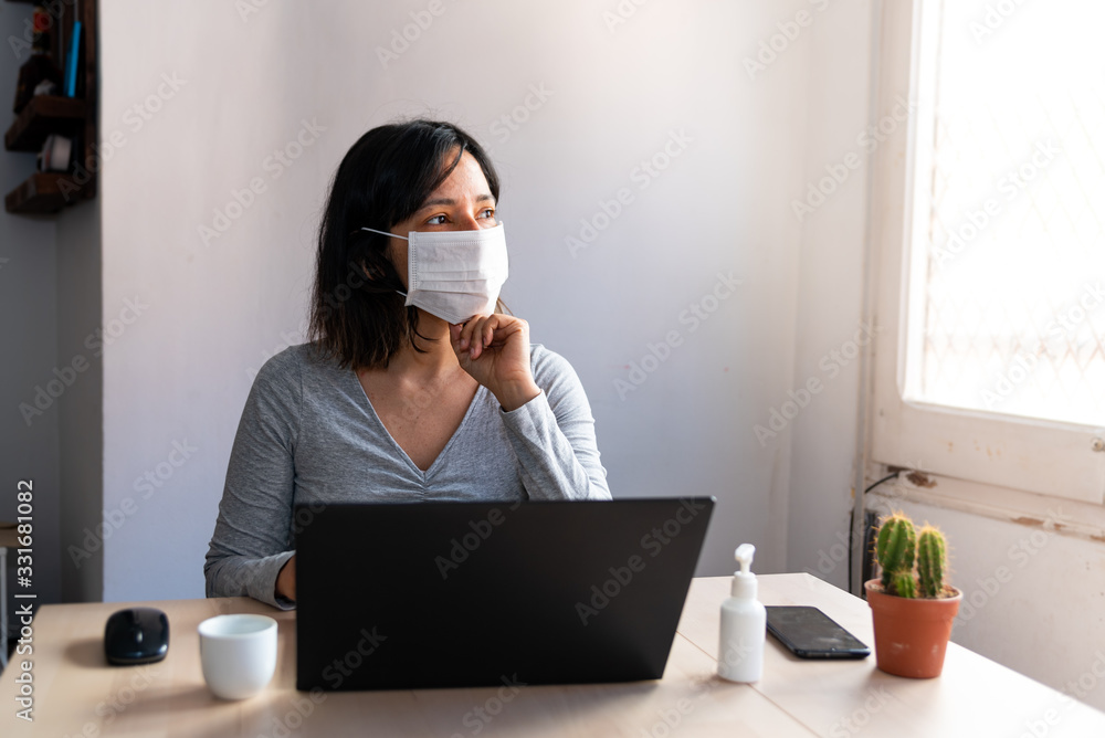 young woman wearing protective face mask and with hand sanitizer in home office looking worried and stressed out of the window due to corona virus - obrazy, fototapety, plakaty 
