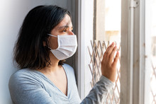 Portrait Of Young Woman Infected With Corona Virus With Face Mask Looking Out Of Window While In Quarantine With Sad And Worried Expression Due To Isolation