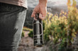 © magdal3na - Men hand with black bottle of wine and vineyard blurred background.