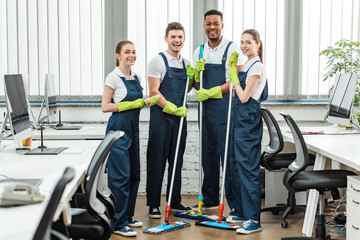 Wall Mural - cheerful multicultural team of cleaners looking at camera while standing with mops in office