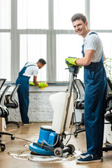 Wall Mural - smiling cleaner washing floor with cleaning machine near colleague wiping window