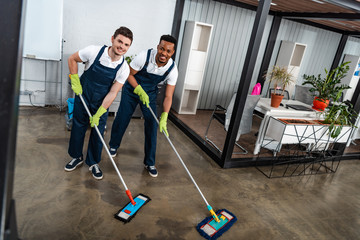 two smiling multicultural cleaners washing floor with mops and looking at camera