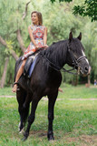Fototapeta Konie - Young woman in a bright colorful dress riding a black horse