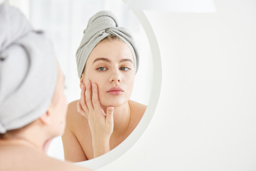 portrait of young girl with towel on head in white bathroom looks and touches her face in the mirror