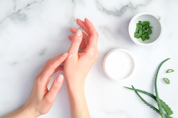 Wall Mural - cosmetic cream on female hands, jar with aloe vera cream and green sliced stems on marble background