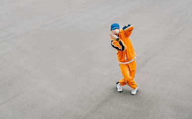 Stylish young man dancing hip hop on an asphalt square, wearing orange clothes. Fashionable street dancer shows hip hop performance on gray asphalt background. Copy space. Background.